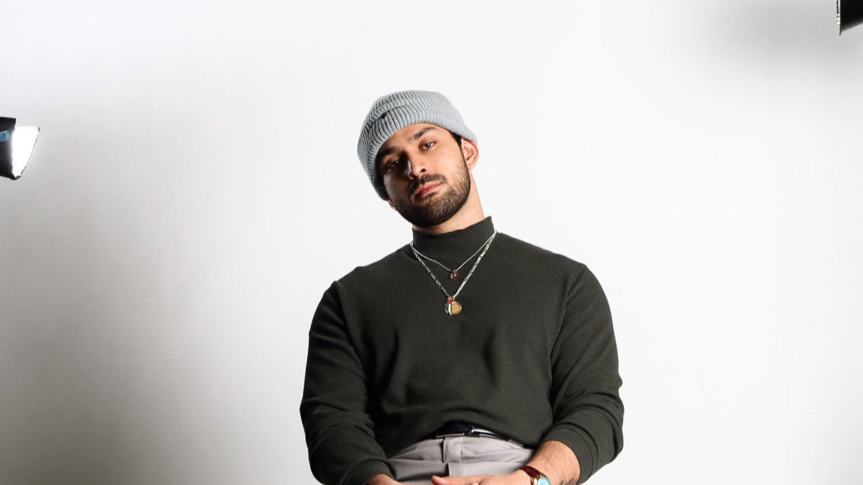 Man sitting on a stool wearing a black sweater, gray pants, and a gray beanie in a studio setting.